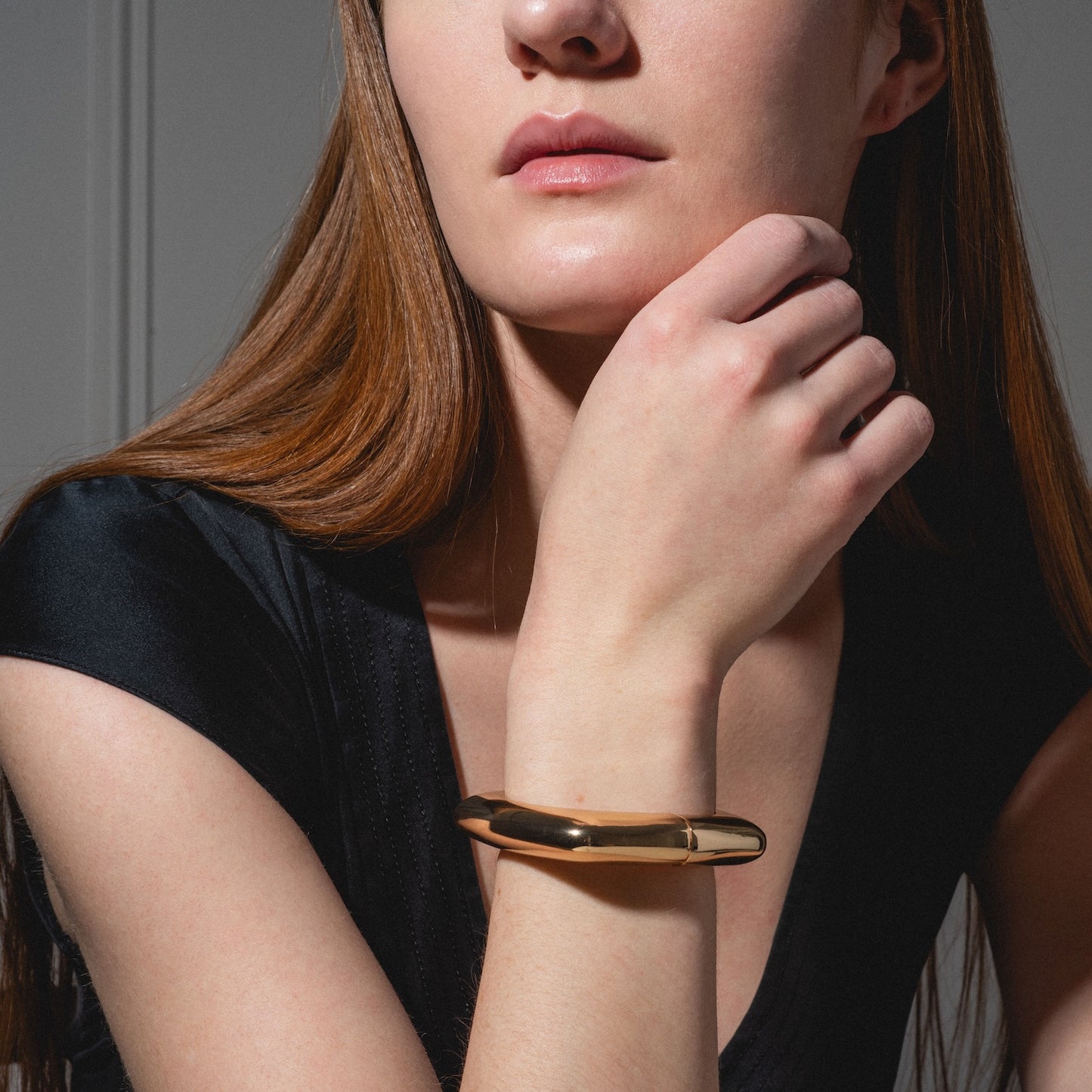 A woman with long auburn hair wears a short-sleeved black top and the Square Cuff Bracelet on her wrist, resting her hand near her chin in soft lighting.