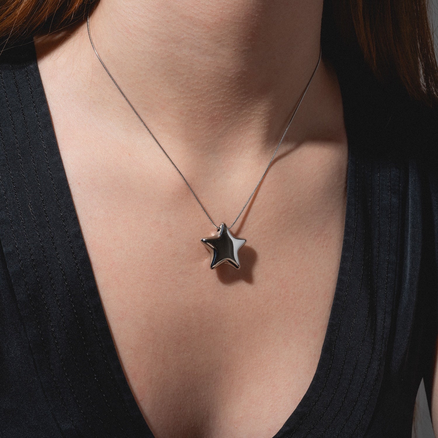 A person wearing a black V-neck top and the Prism Necklace, featuring a shiny metallic star-shaped pendant. The image focuses on the pendant and the person's upper chest.