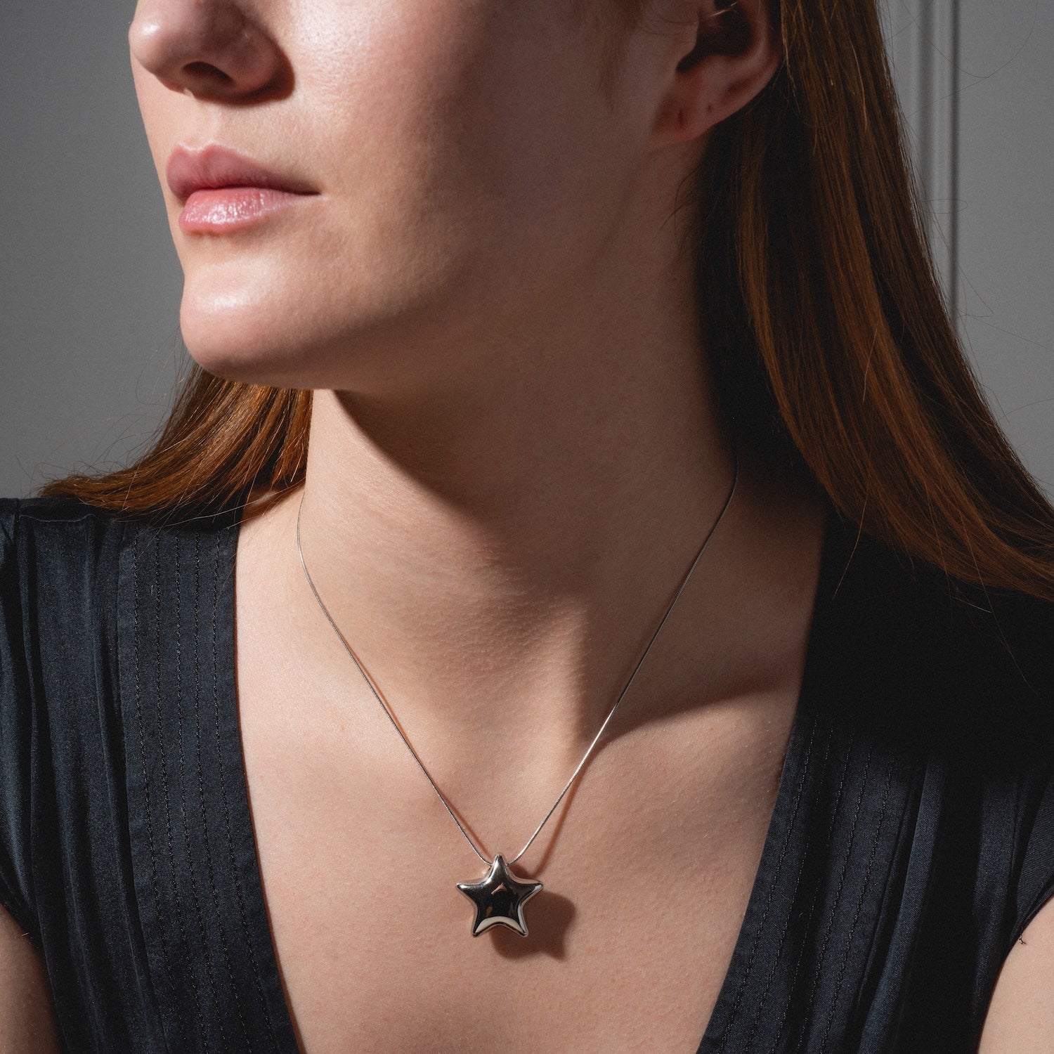 A woman with long reddish hair, wearing a black top and a silver Prism Necklace with a shiny star pendant, shown from the shoulders up against a neutral background.