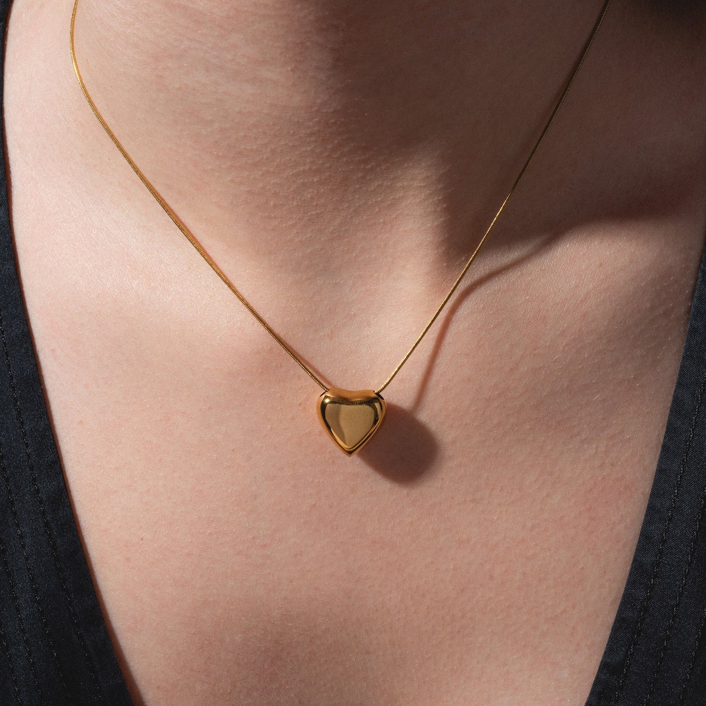 A close-up of a person's neck wearing the Yellow Necklace, a delicate gold piece featuring a small, shiny heart-shaped pendant. The person is dressed in a black V-neck top.