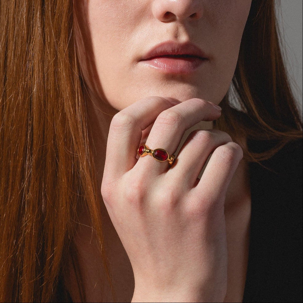 A woman with long red hair and fair skin gazes at the camera, resting her chin on her hand adorned with the Bita Ring. She wears a black top against a softly lit, neutral background.