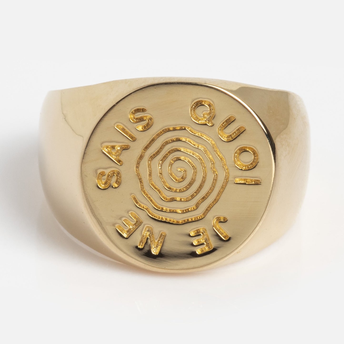 The I Like It Here Club x Local Eclectic Je Ne Sais Quoi Pinky Ring is a gold ring with a flat circular face engraved with a spiral and the words JE NE SAIS QUOI in capital letters around it.