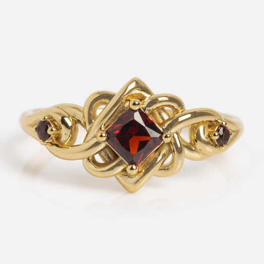 The Lover's Knot Ring is a gold ring with an intricate, interwoven design featuring a square-cut deep red center gemstone flanked by two small round red stones on each side.