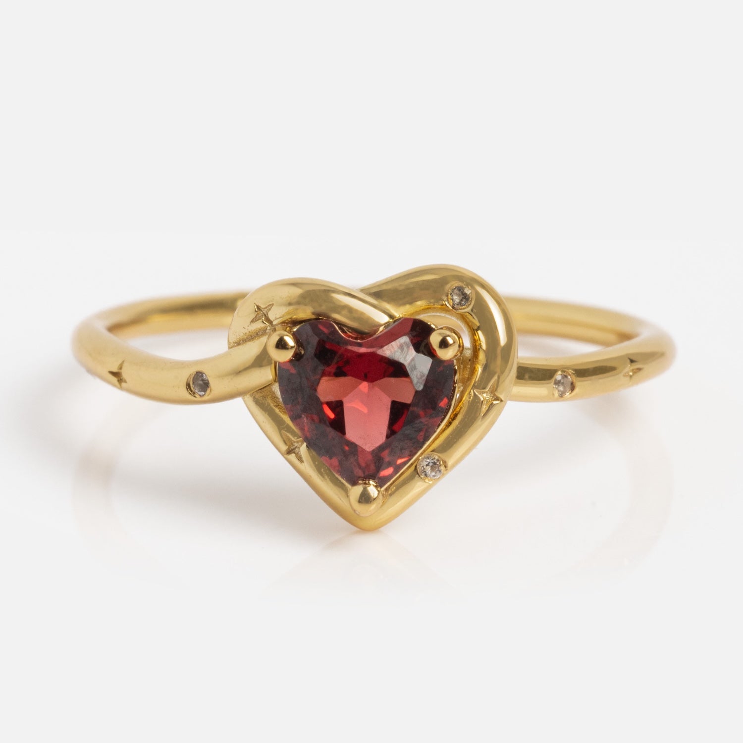 The Lover's Knot Heart Ring features a heart-shaped red gemstone at its center, encircled by a gold heart frame with small clear stones, all set against a white background.