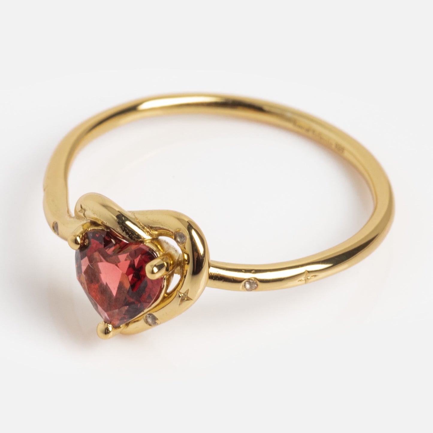 The Lover's Knot Heart Ring showcases a heart-shaped red gemstone at its center, accented by star engravings and tiny diamonds along the delicate gold band. Its elegant design stands out against a plain white background.