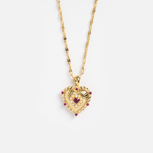 The Til The End and After Lover's Locket Necklace is a gold necklace with a heart-shaped pendant, featuring intricate detailing and small pink gemstones, shown on a plain white background.