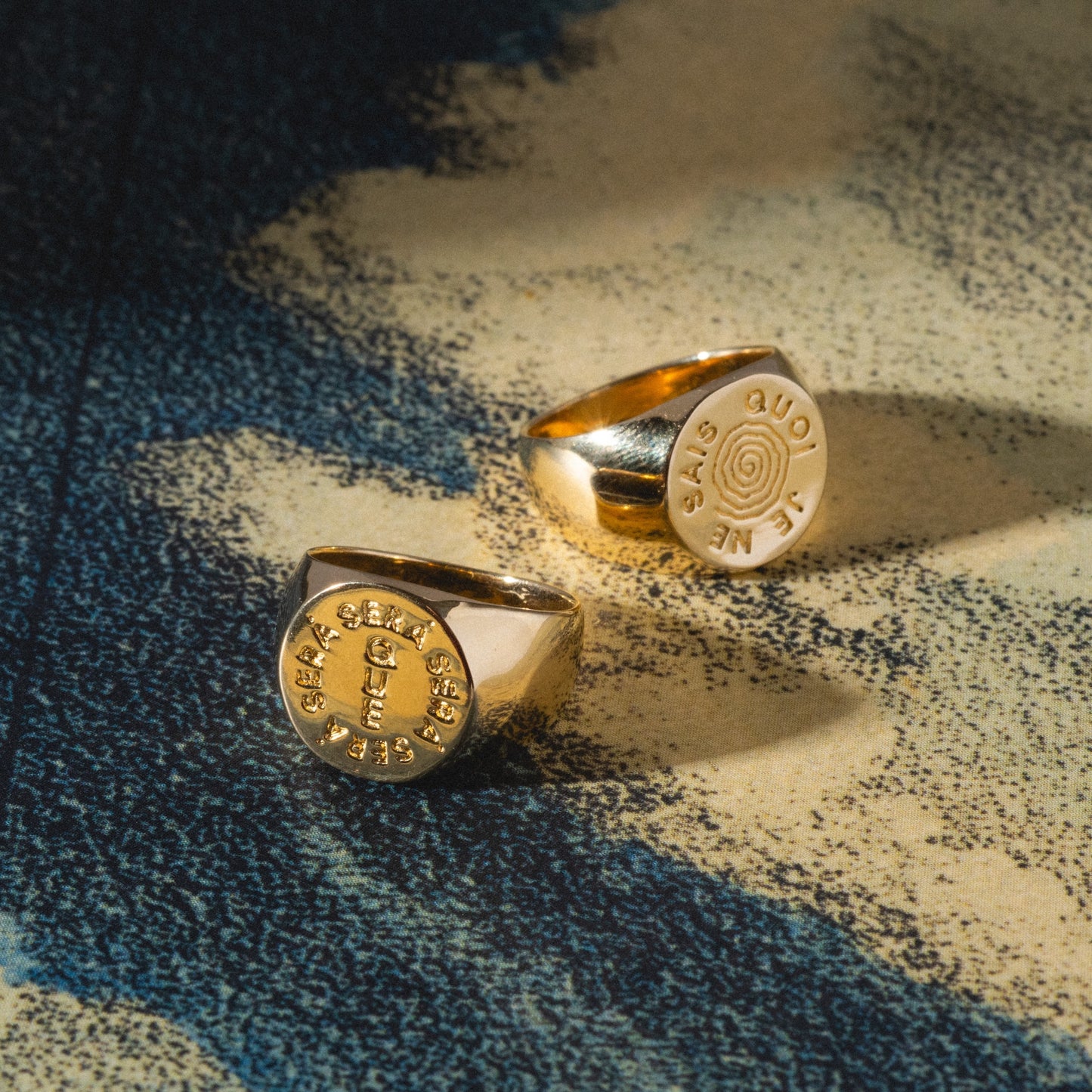 The I Like It Here Club x Local Eclectic Je Ne Sais Quoi Pinky Rings rest on a textured blue and beige surface, their round, flat faces engraved with text and symbols, the lighting accentuating their lustrous finish.