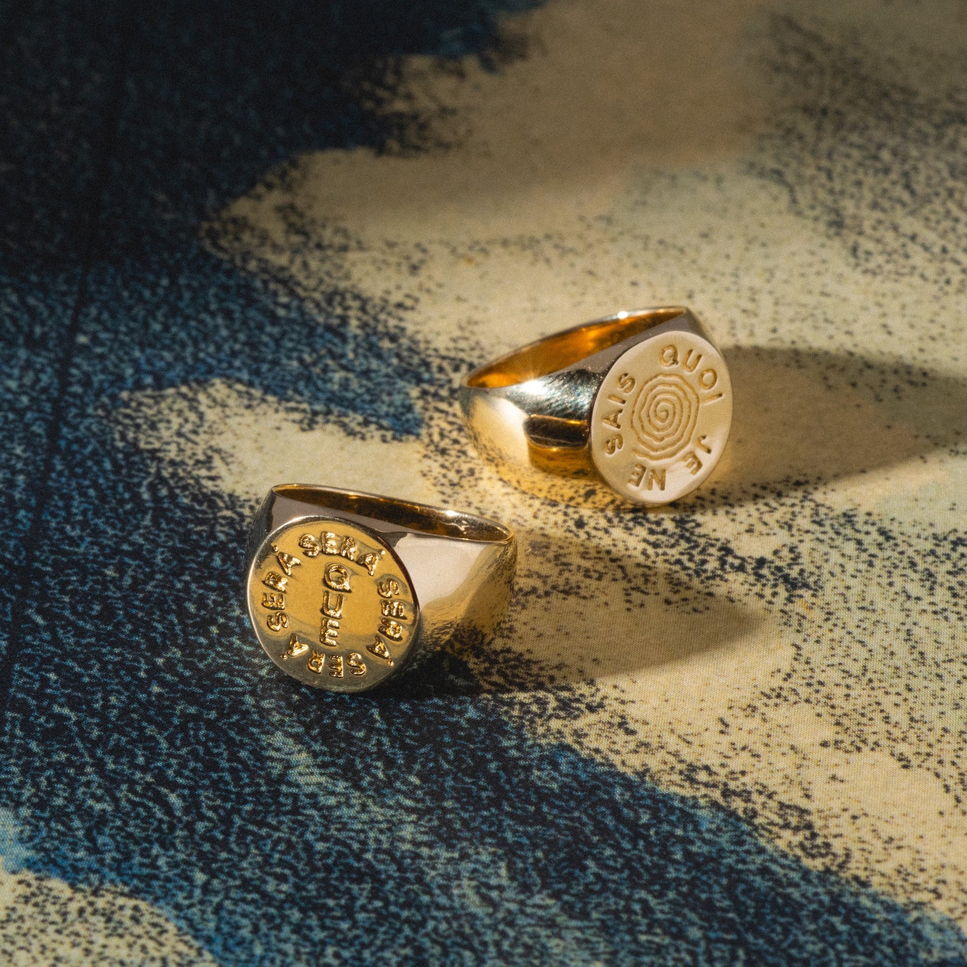 The I Like It Here Club x Local Eclectic Je Ne Sais Quoi Pinky Rings rest on a textured blue and beige surface, their round, flat faces engraved with text and symbols, the lighting accentuating their lustrous finish.