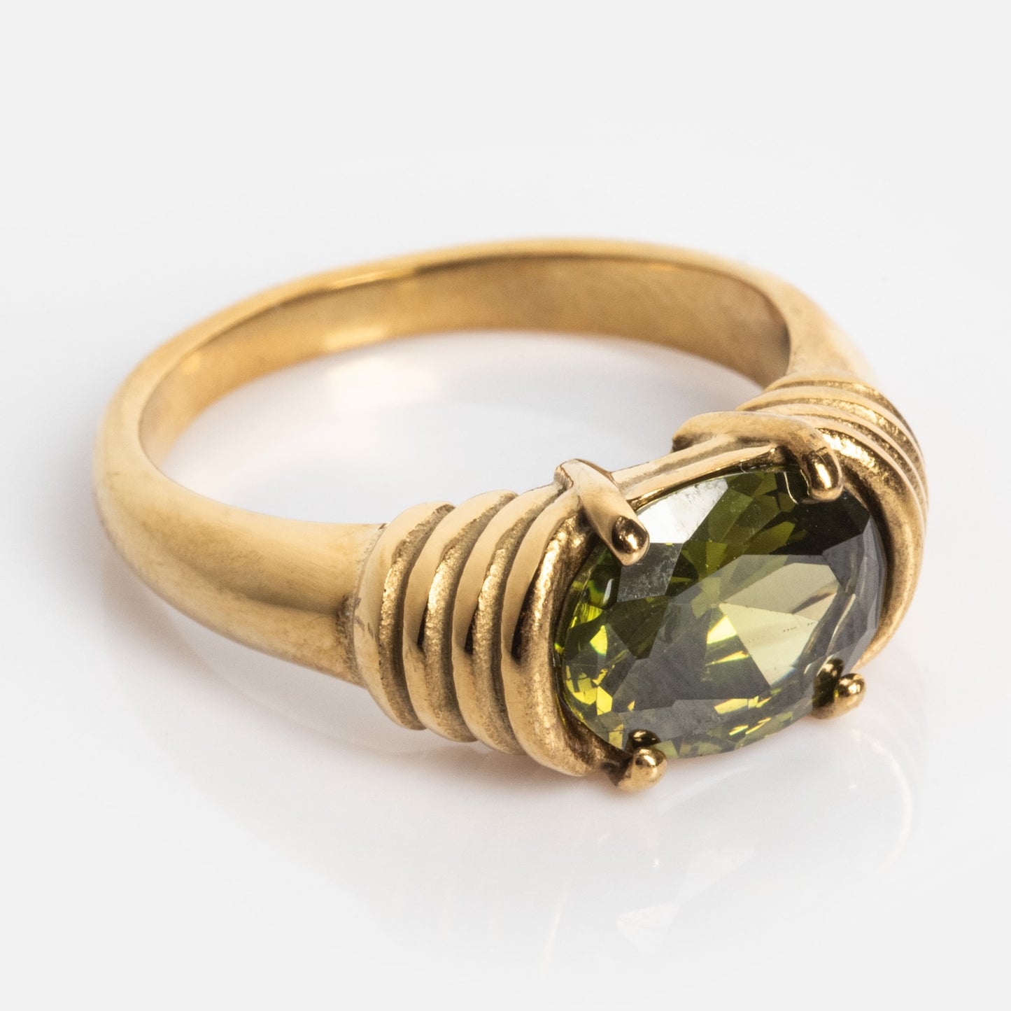 The Stone Statement Ring showcases a large, oval-cut green gemstone set in a gold wire-wrapped band, elegantly displayed on a reflective white surface.