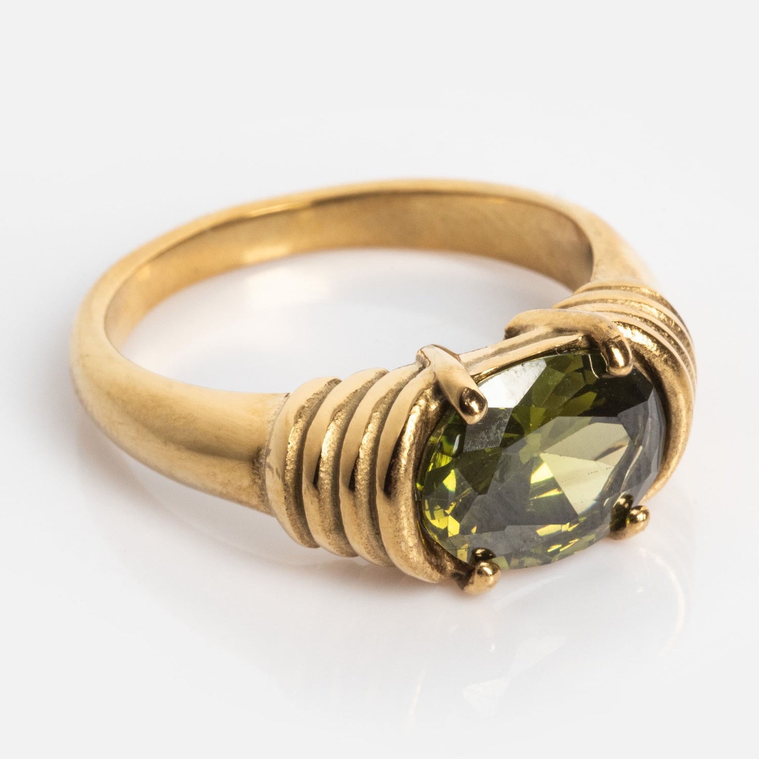 The Stone Statement Ring showcases a large, oval-cut green gemstone set in a gold wire-wrapped band, elegantly displayed on a reflective white surface.