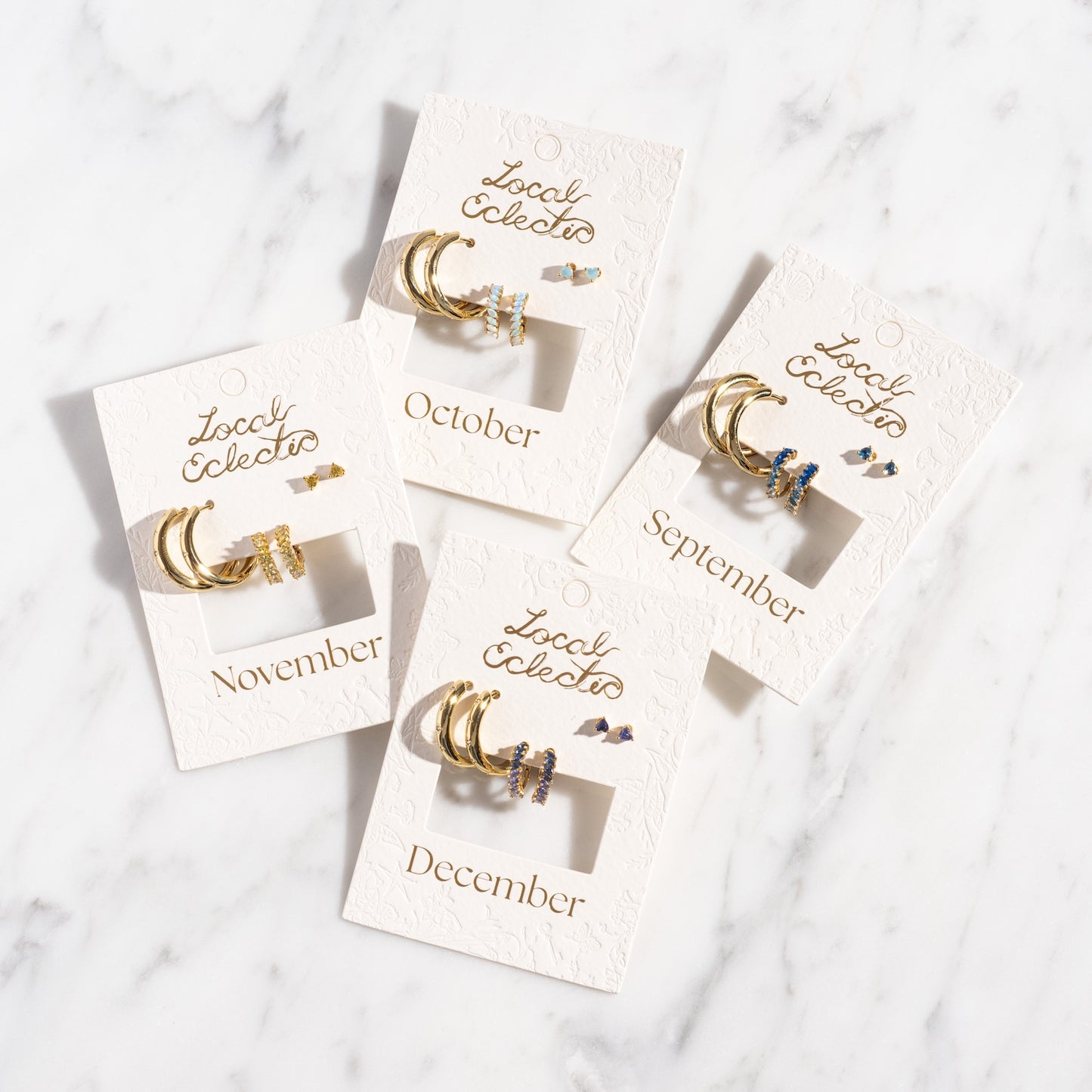 Four Bold Birthstone Earring Sets with gemstone accents, each on a white card labeled September, October, November, or December, are displayed on a white marble surface.