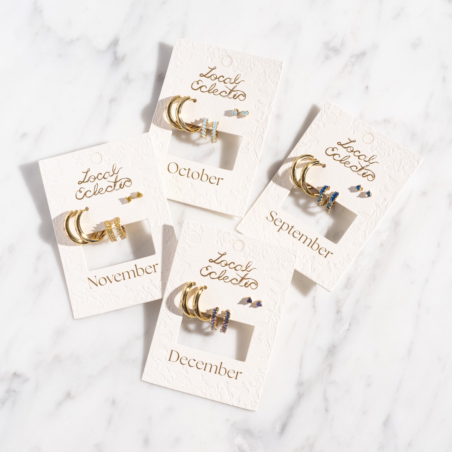 Four Bold Birthstone Earring Sets with gemstone accents, each on a white card labeled September, October, November, or December, are displayed on a white marble surface.