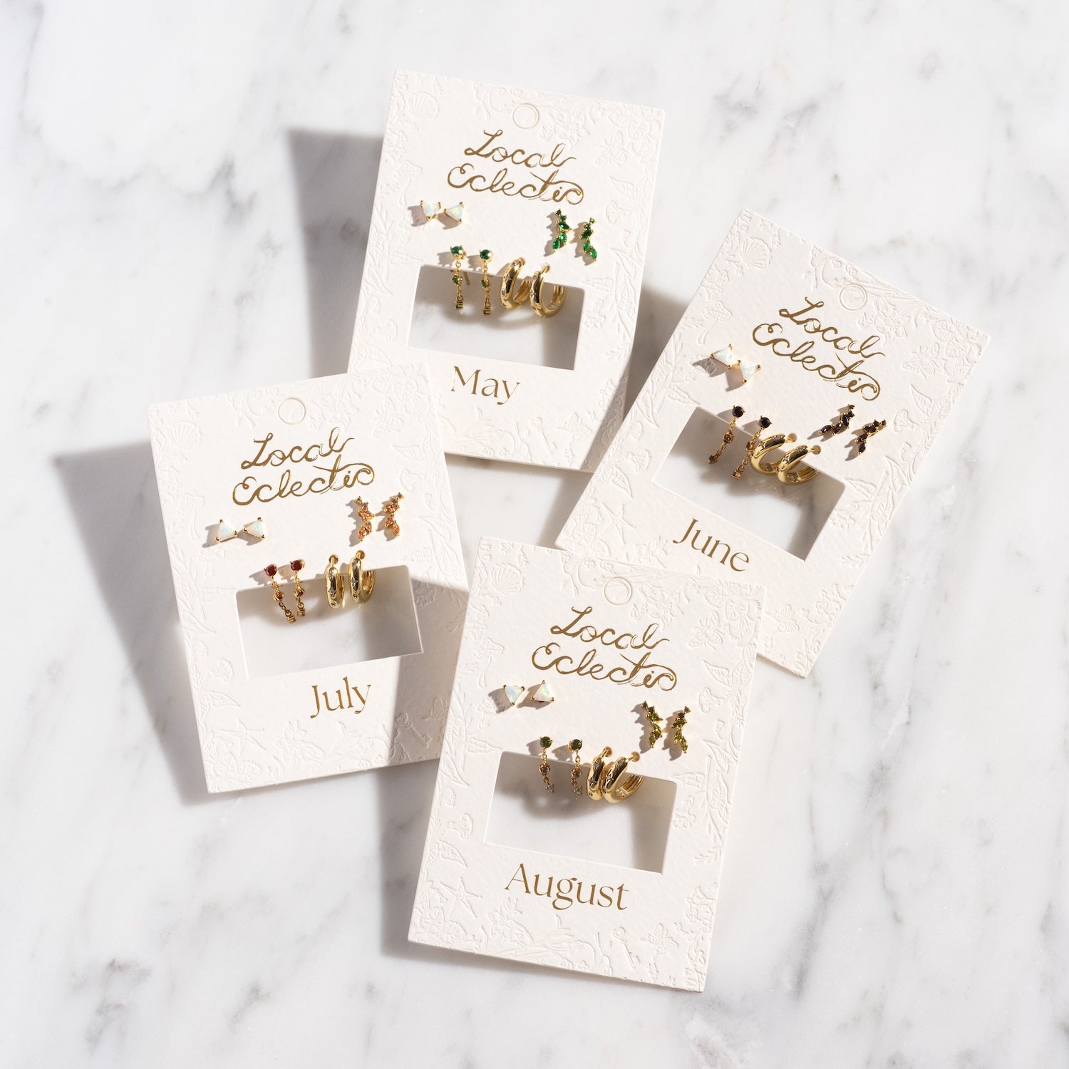 Four jewelry cards labeled May, June, July, and August showcase the Classic Birthstone Earring Set—14k gold plated studs—on a white marble surface with embossed designs and gold "Local Eclectics" lettering.