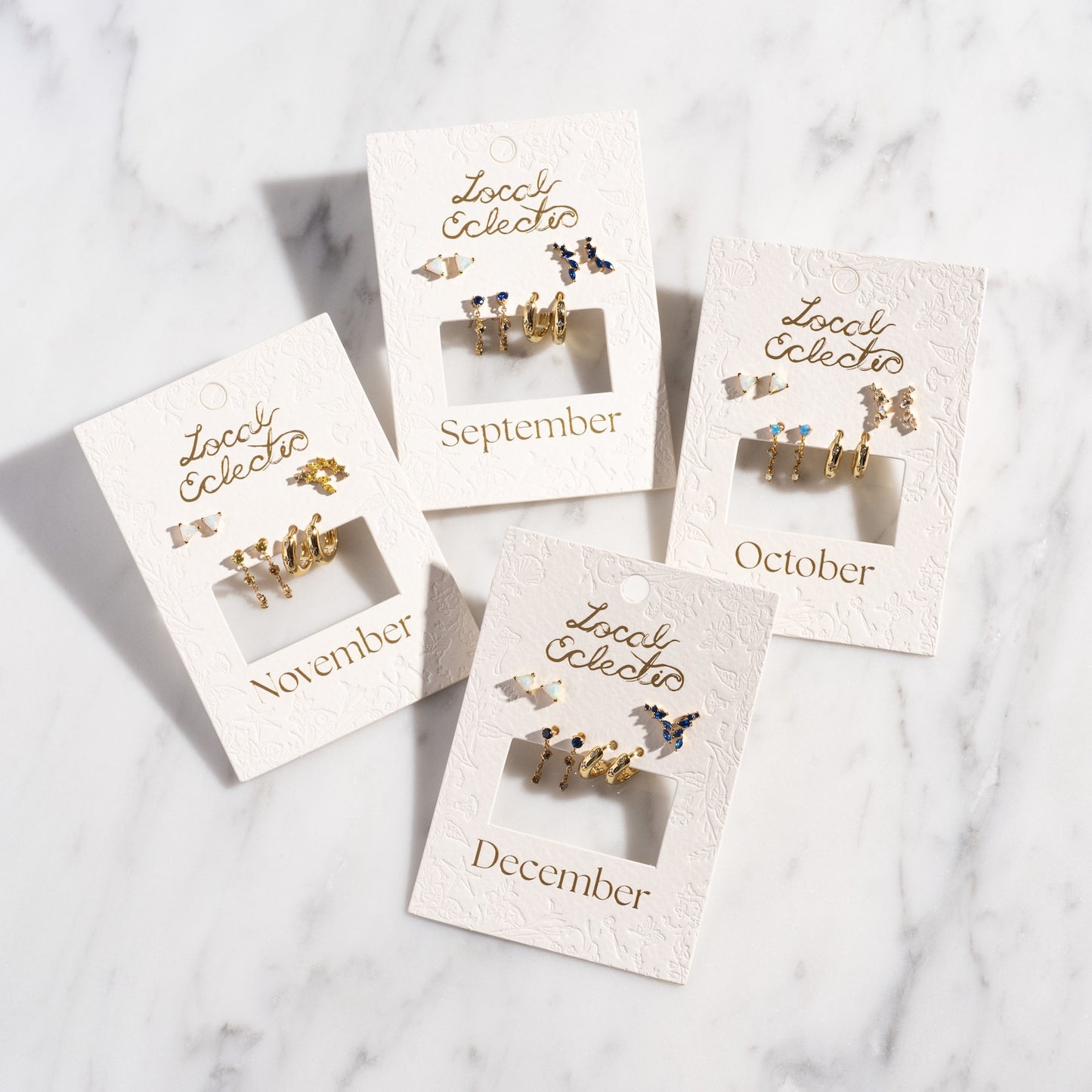 Four Classic Birthstone Earring Sets, plated in 14k gold and labeled for September through December, perfect for a personalized ear stack, displayed on embossed Local Eclectic cards atop a white marble surface.
