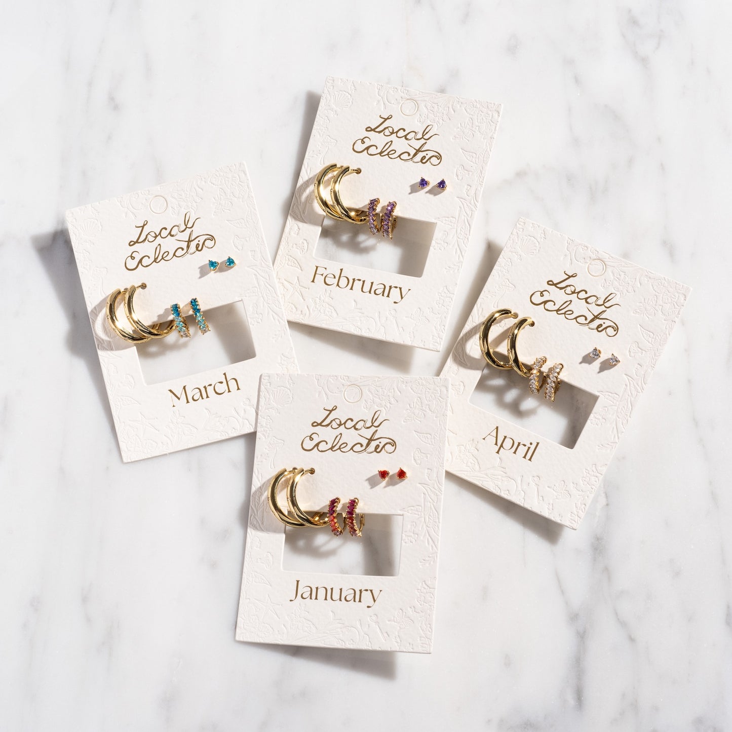 The Bold Birthstone Earring Set features gold plated birthstone earrings for January, February, March, and April with colorful gemstones, beautifully arranged on a marble surface.