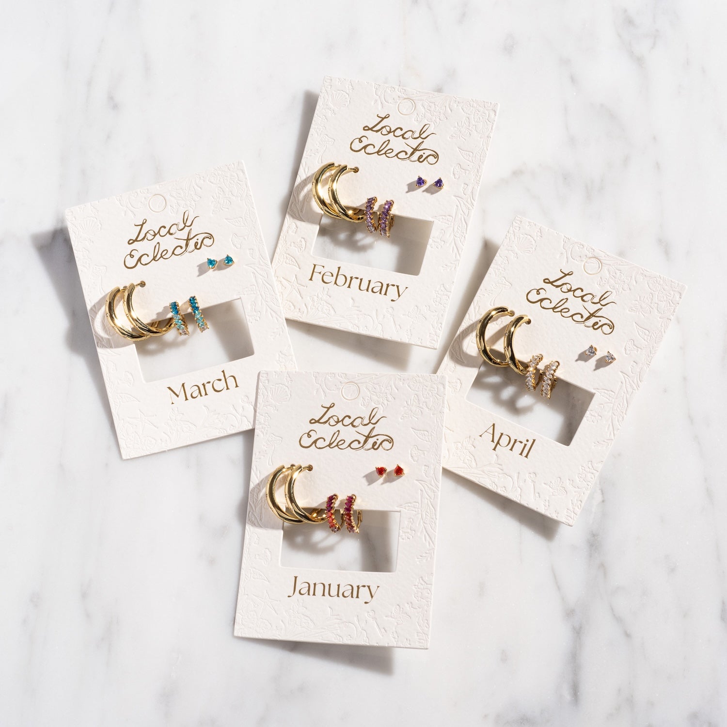 The Bold Birthstone Earring Set features gold plated birthstone earrings for January, February, March, and April with colorful gemstones, beautifully arranged on a marble surface.