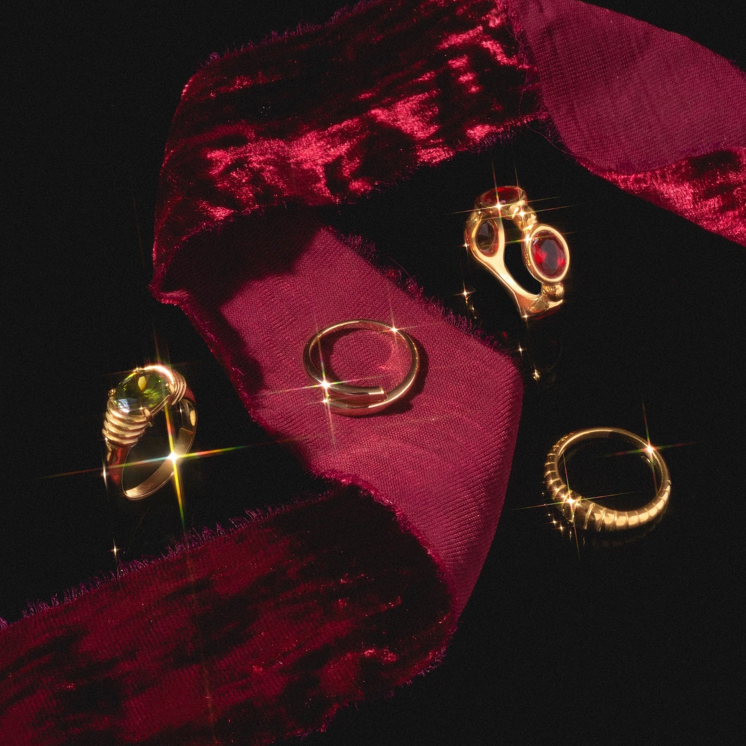 The Wrap Around Ring is displayed on a dark surface with three other gold rings and a deep red velvet ribbon, their gemstones sparkling in the light.