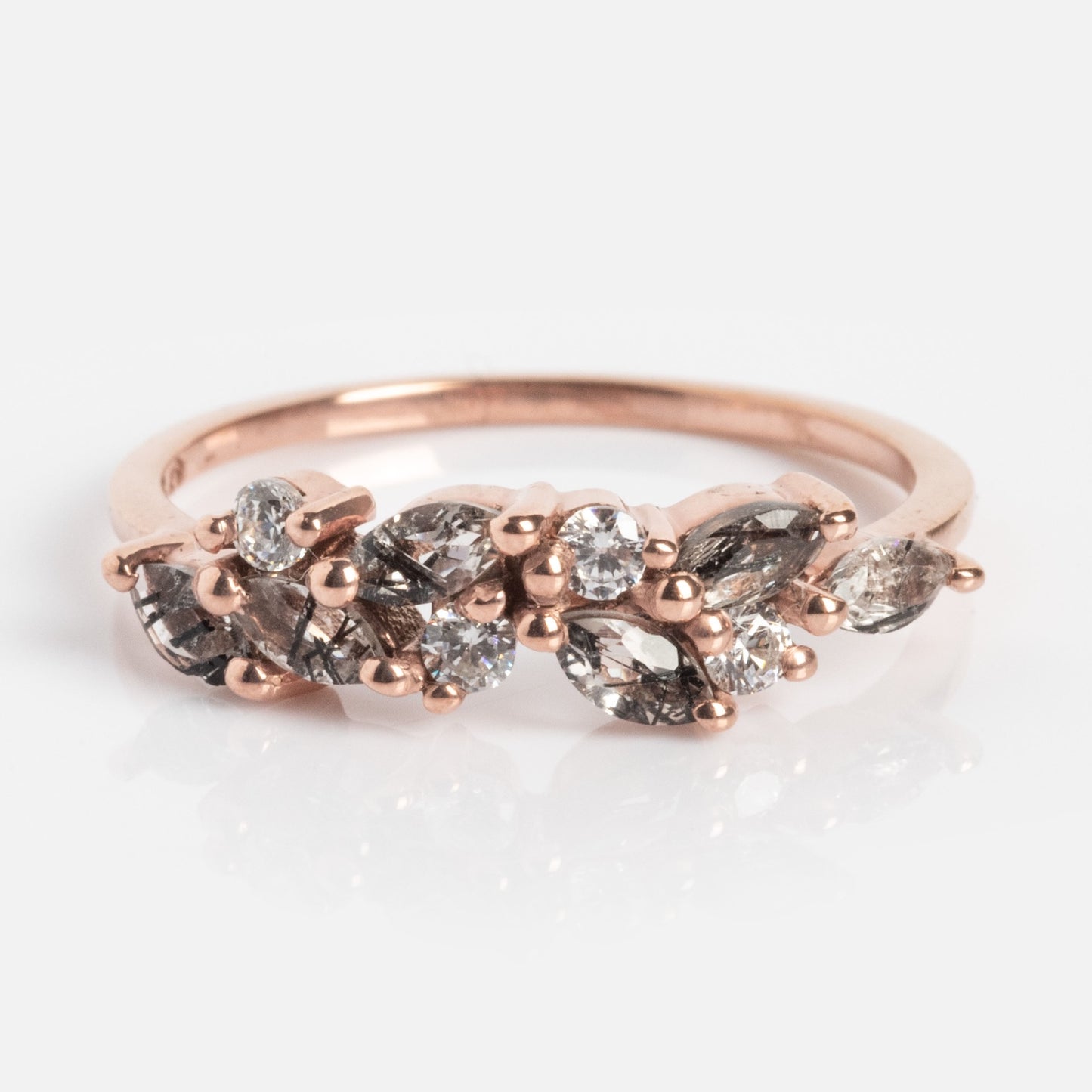 The Gemstone Orchard Ring is a rose gold band with a curved cluster of marquise and round-cut clear, gray gemstones, and moss agate in an organic pattern, shown against a white background.