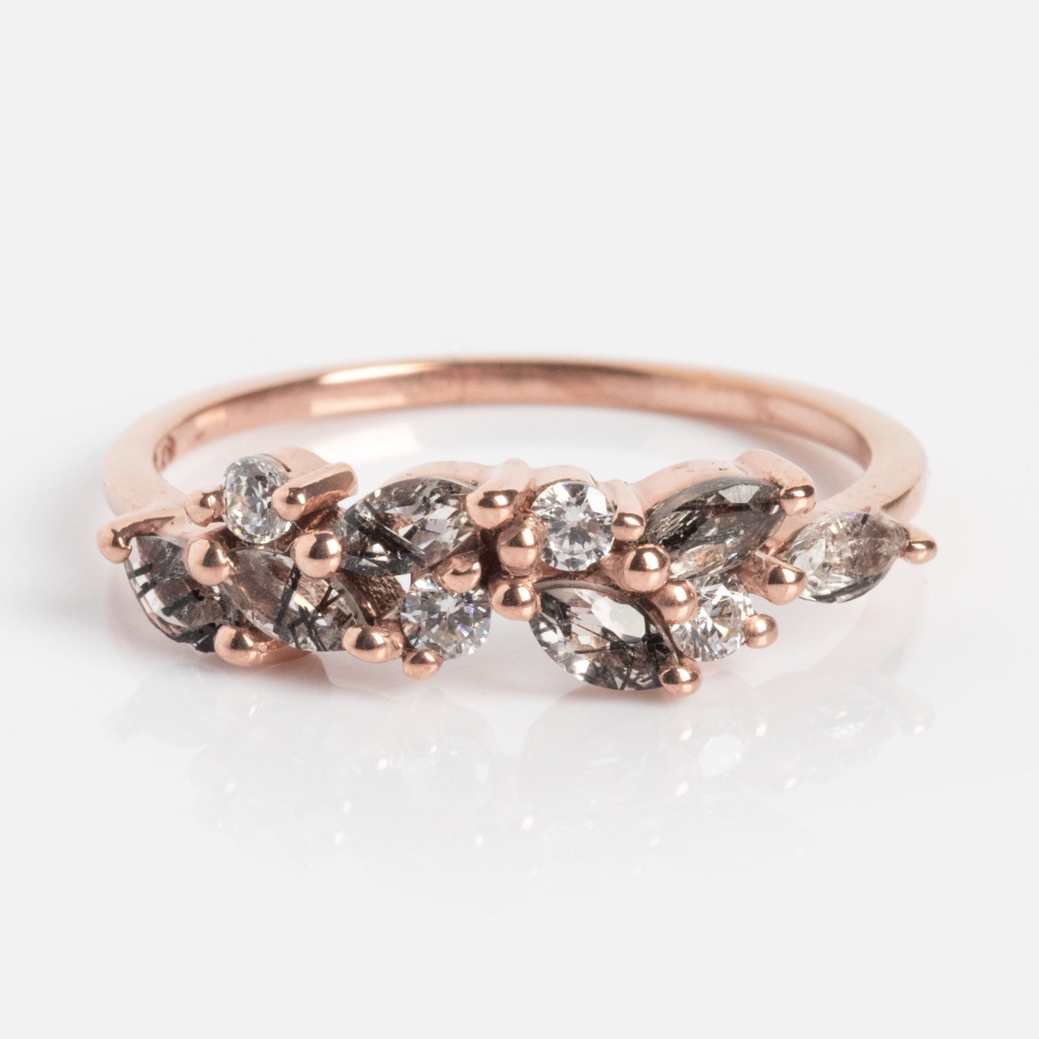 The Gemstone Orchard Ring is a rose gold band with a curved cluster of marquise and round-cut clear, gray gemstones, and moss agate in an organic pattern, shown against a white background.