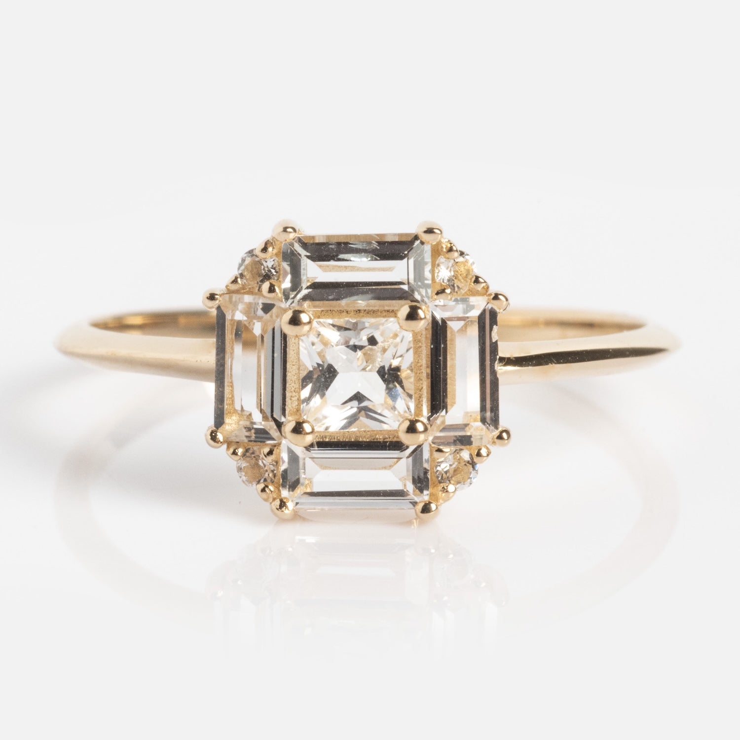The 14k Vintage Inspired Art Deco Engagement Ring showcases a square clear gemstone with beveled edges in a geometric prong setting, surrounded by sparkling accent stones and crafted from recycled gold.