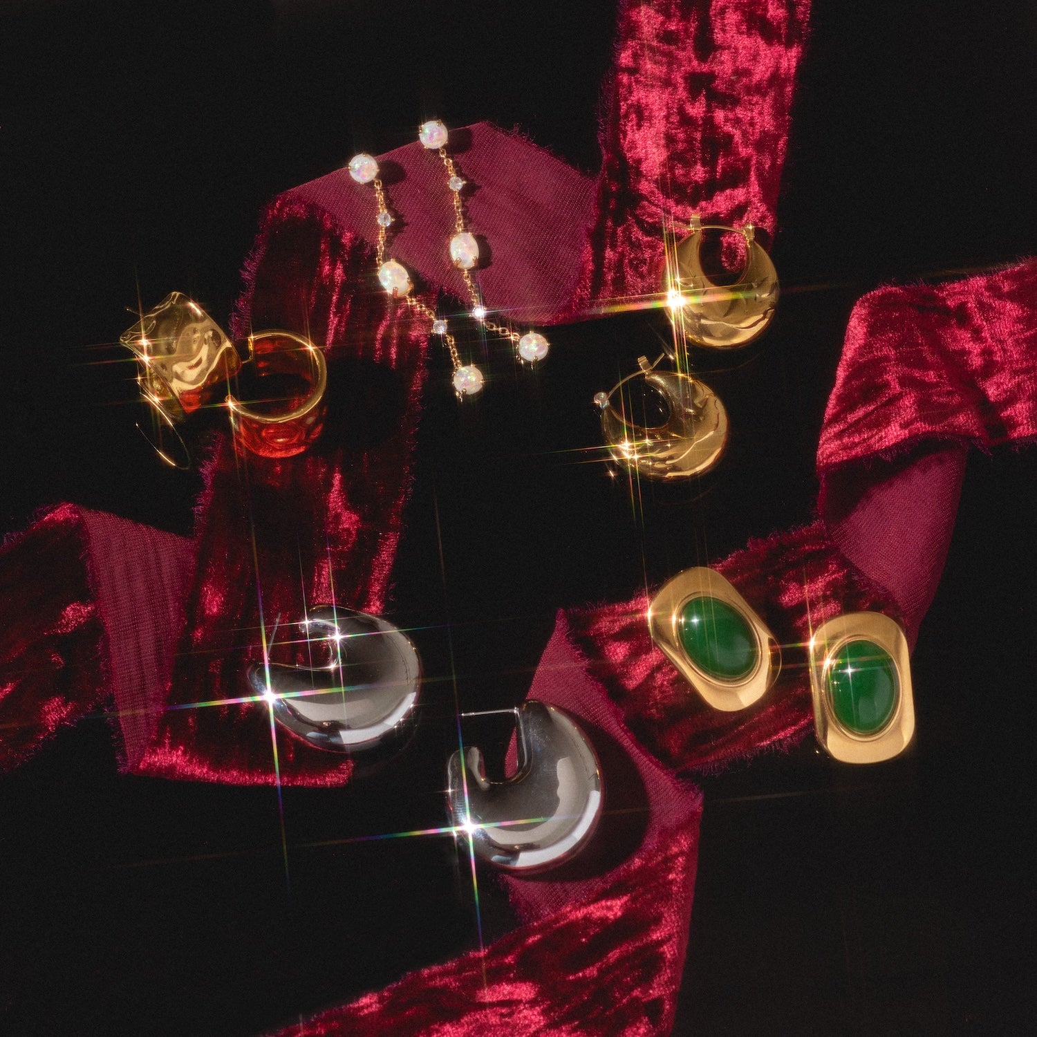 Ashley Earrings in gold, silver, and green gemstone styles displayed on a dark surface with a burgundy velvet ribbon, sparkling reflections highlighting the jewelry.