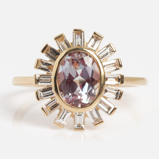 The 14k Alexandrite and Diamond Sun Ring features an oval-cut purple alexandrite center, encircled by a sunburst halo of baguette-cut diamonds, all set on a slim gold band against a white background.