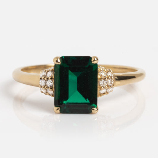The 14k Classic Emerald and Diamond Cocktail Ring features a rectangular emerald-green center stone flanked by clusters of small round diamonds, shown on a plain white background.