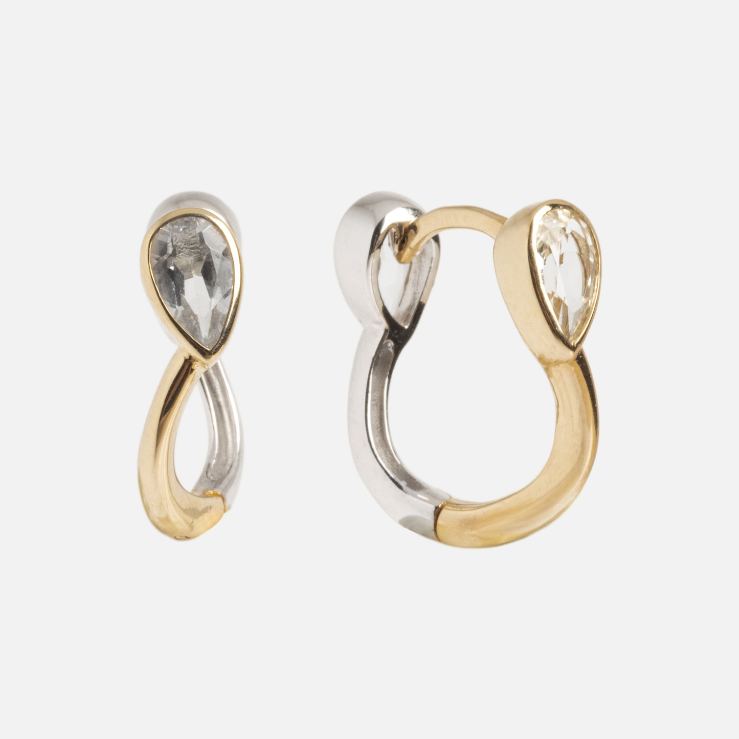The Solid Gold Mixed Metal Duo Huggies are earrings with intertwined gold and silver tones in a loop, featuring teardrop-shaped clear gemstones, displayed on a white background.
