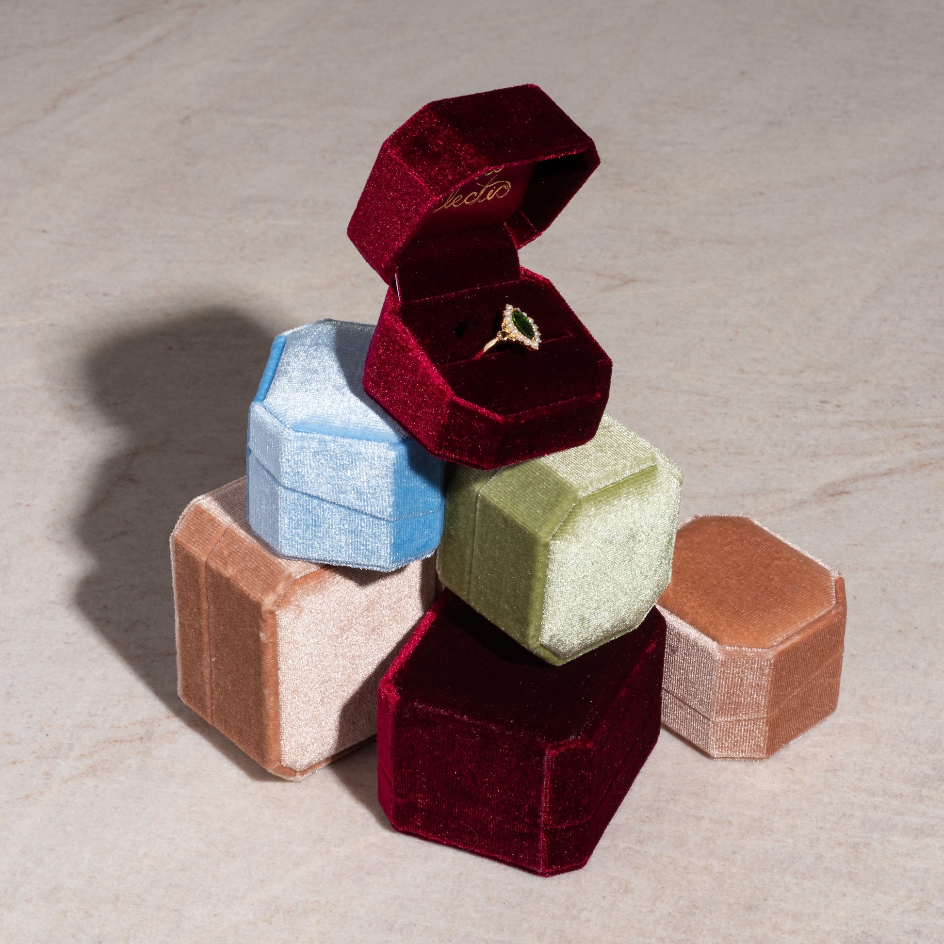 A gold ring with a green gemstone rests in an open Fancy Velvet Ring Box, stacked on closed velvet boxes in blue, green, pink, and tan atop a light-colored surface.