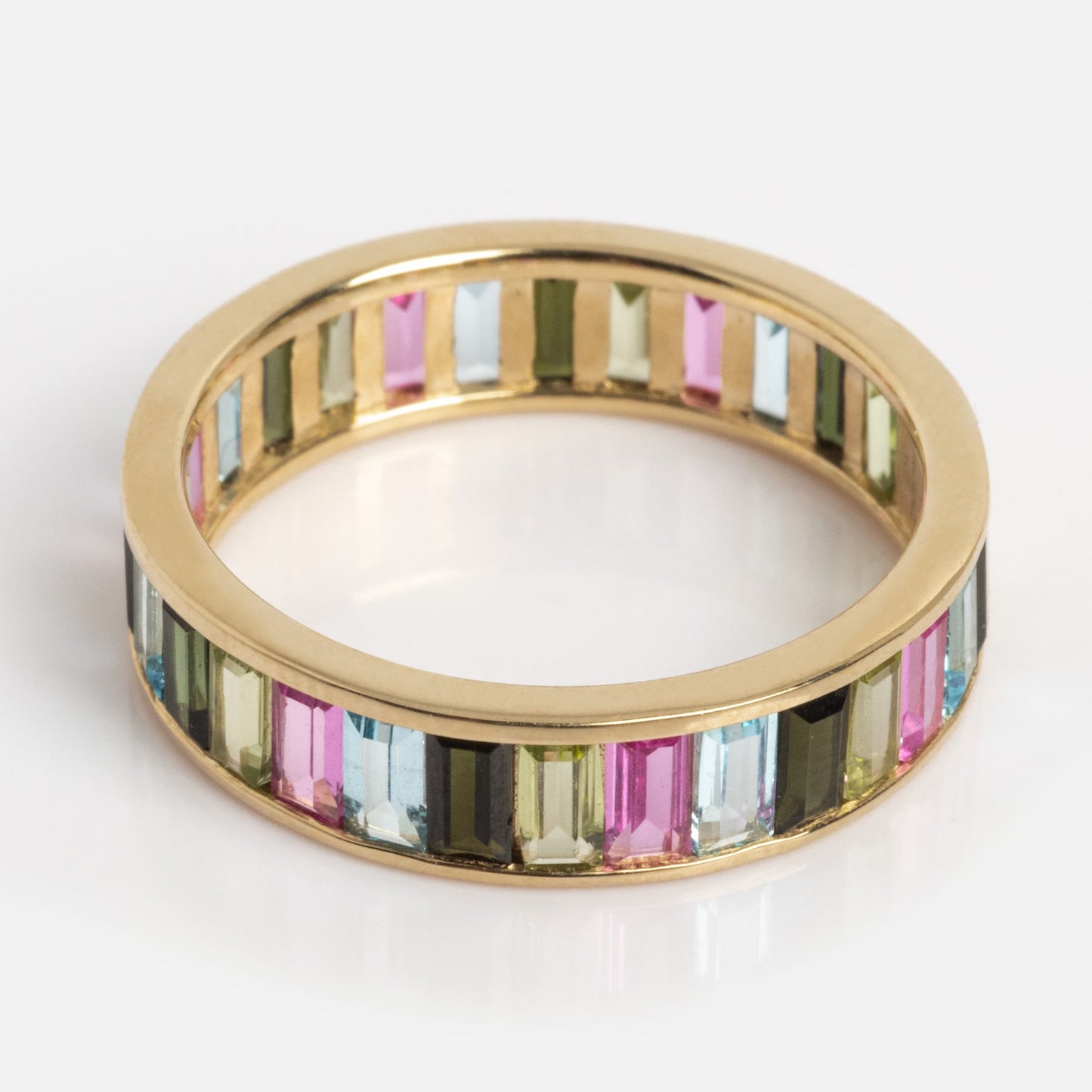 The Solid Gold Gemstone Kaleidoscope Eternity Band features rectangular multicolored gemstones set in a continuous row around the gold band, shown against a white background.