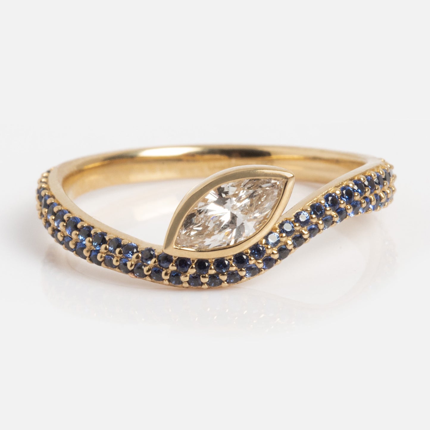 The Solid Gold Nesting Gleam Sapphire and Diamond Ring showcases a marquise-cut diamond center on a gold band accented with blue sapphires in a wave design, displayed against a white background.