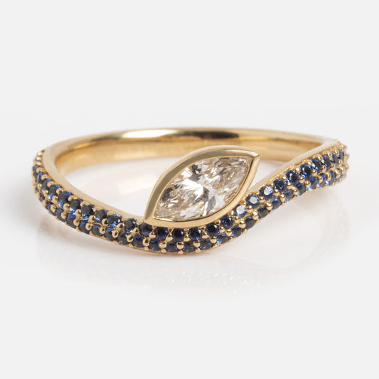 The Solid Gold Nesting Gleam Sapphire and Diamond Ring showcases a marquise-cut diamond center on a gold band accented with blue sapphires in a wave design, displayed against a white background.