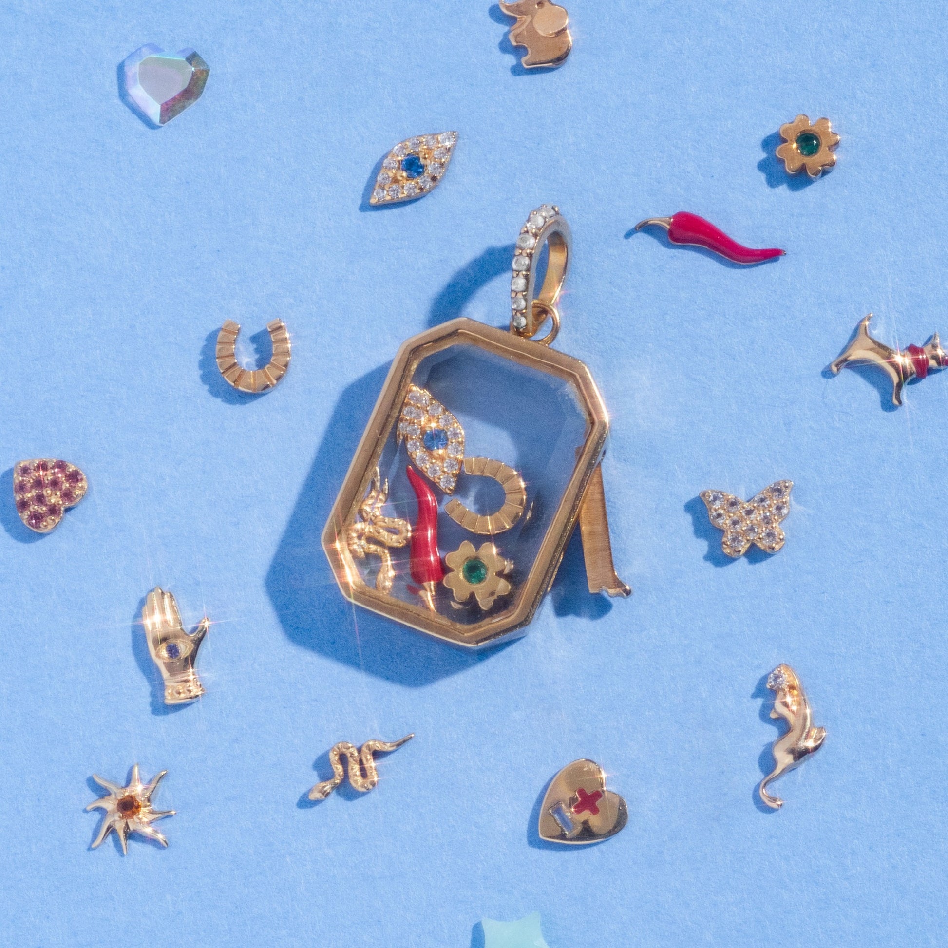 A Solid Gold Tiny Lucky Charm for Glass Charm Locket, featuring yellow gold charms like a snake, flower, and red horn, rests on a light blue surface with small hand, eye, star, heart, and other decorative good luck symbols around it.