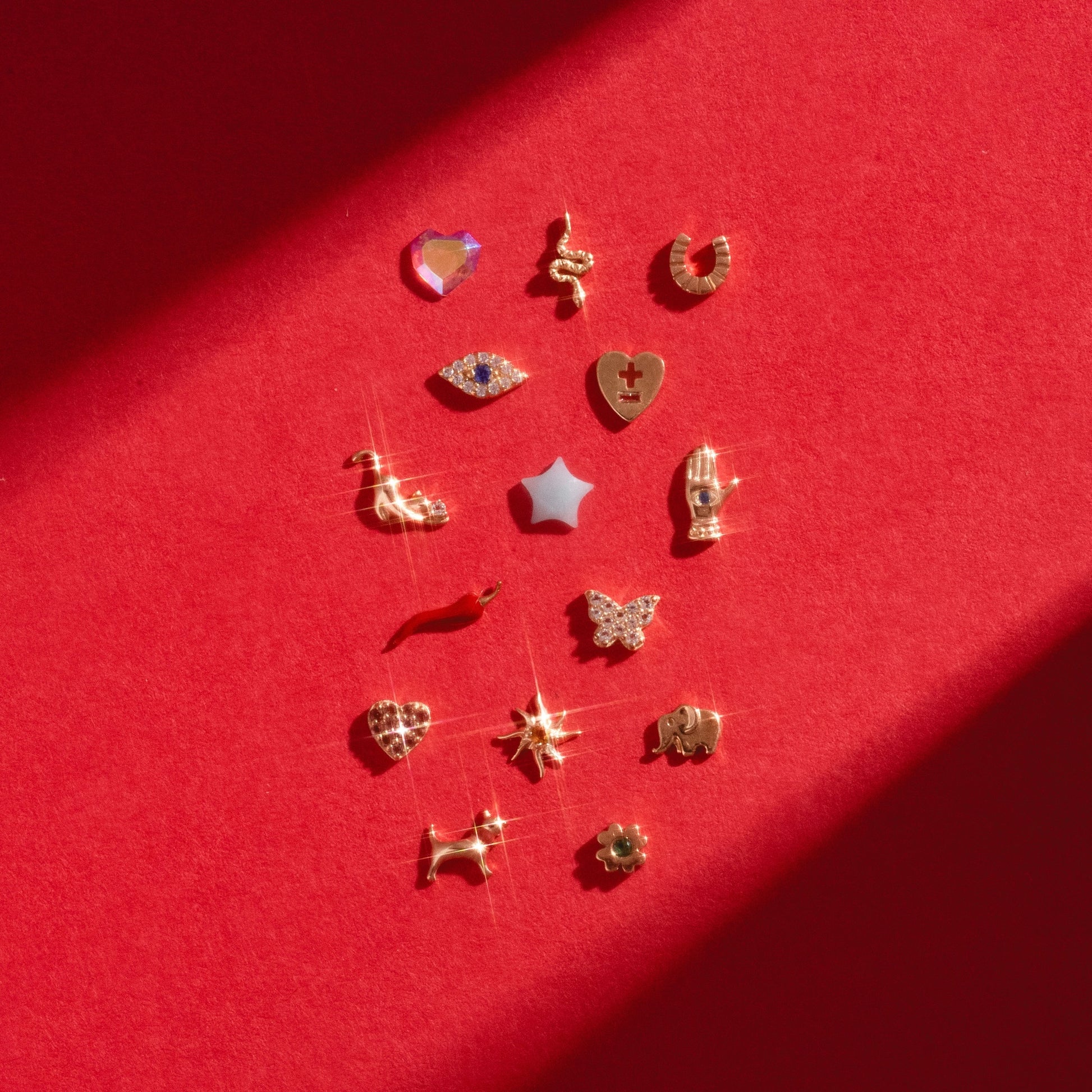 A set of 16 Solid Gold Tiny Lucky Charms for Glass Charm Locket, featuring shapes like heart, star, butterfly, and elephant—displayed in rows on a vibrant red background with sunlight creating charming shadows.