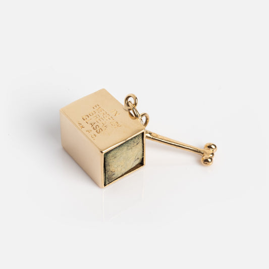 A Vintage 14k Emergency Money Charm, a small gold-toned metal box with an attached key, engraved text on one side, and a metal loop on top, rests on a white surface.