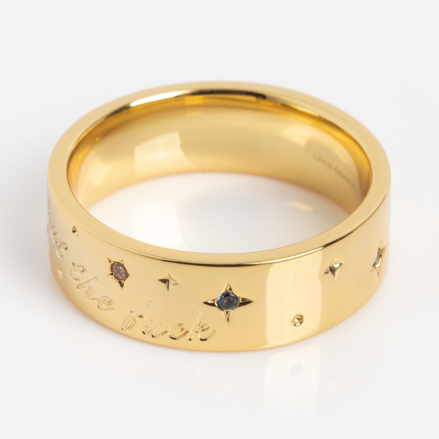 The What the Fuck Cigar Band is a wide gold ring with small star-shaped engravings, colored gemstones set in each star, and a cursive "What the Fuck" inscription along the outer band.