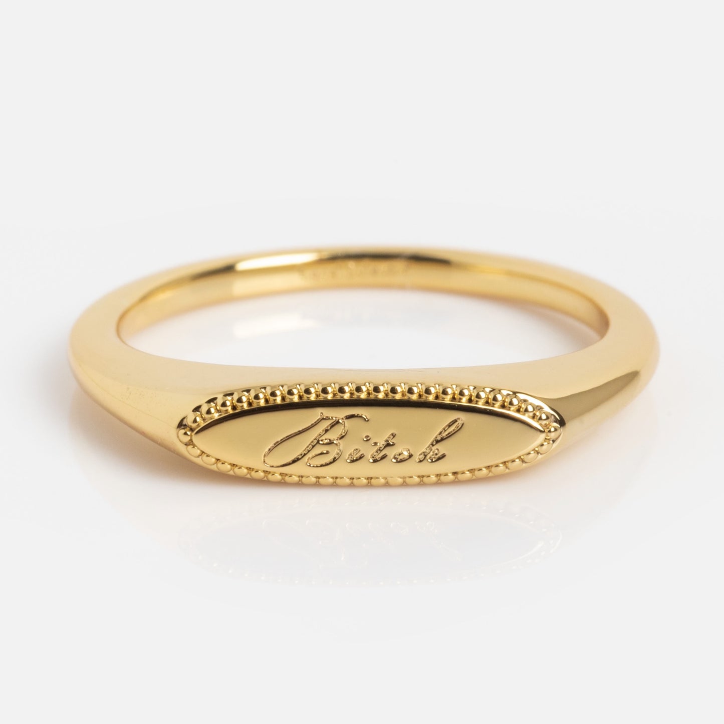The Tiny Bitch Ring is a gold ring with an oval face, "Bitch" engraved in cursive, and a beaded border, displayed on a white background.