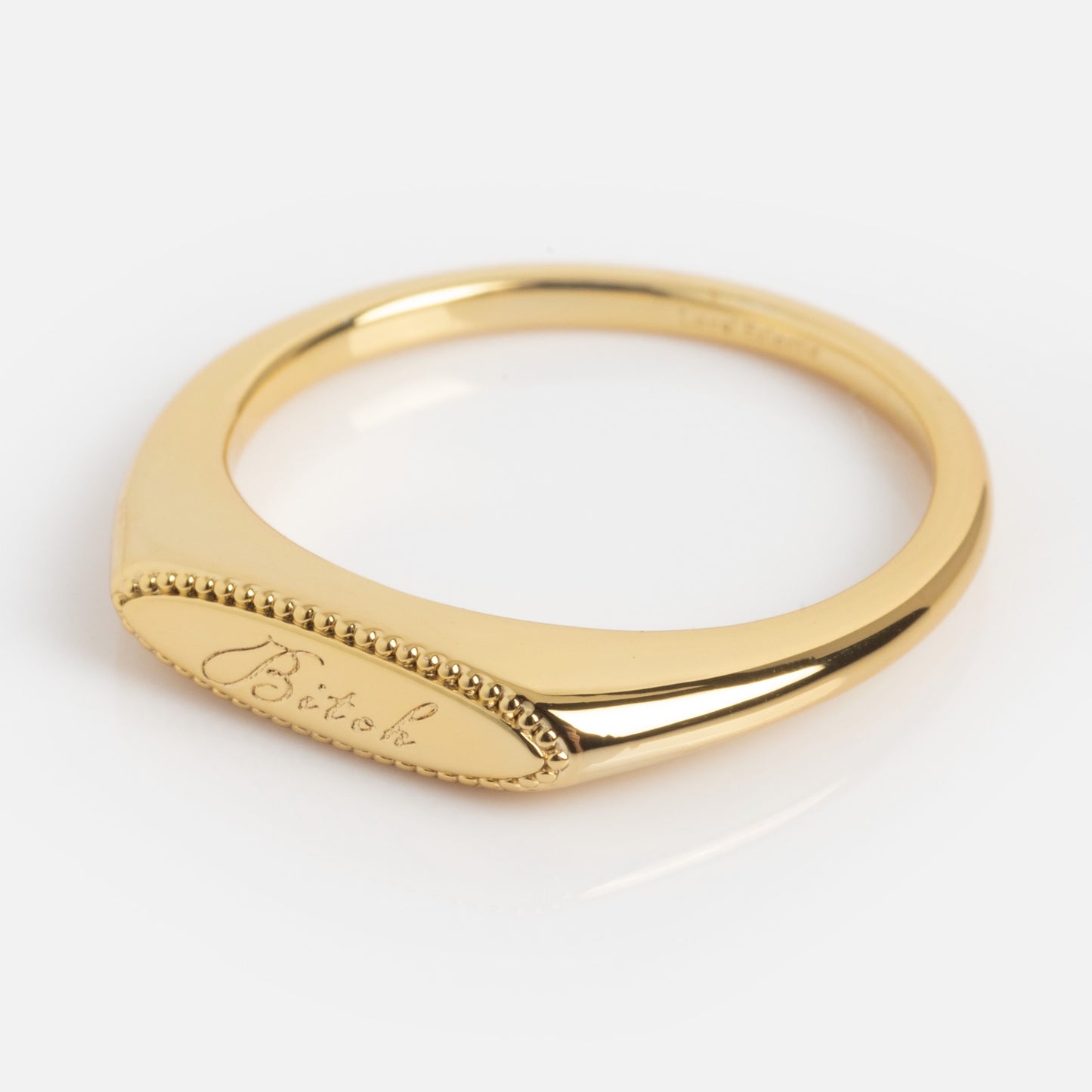 A delicate gold Tiny Bitch Ring featuring a flat, oval top engraved with "Bitch" in cursive and accented by a beaded edge.