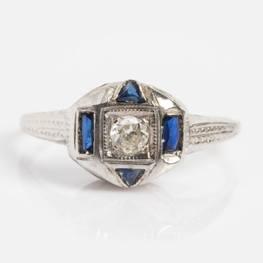 The Vintage 18k Diamond and Sapphire Deco Cluster Ring, size 7, features a round diamond center with rectangular and triangular blue sapphires, accented by detailed engraved designs on the band.