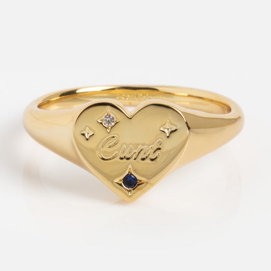 The Tiny Cunt Signet Ring is a gold ring featuring a heart-shaped face engraved with "Cunt," accented by two small gemstones and engraved stars. Its bold, unique design stands out against a plain white background.