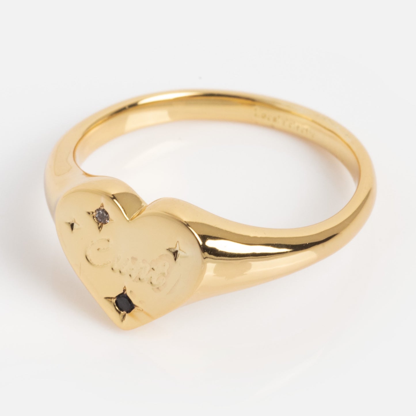 The Tiny Cunt Signet Ring is a gold ring with a heart-shaped face engraved in cursive, featuring small black stones and star accents, shown on a plain white background.