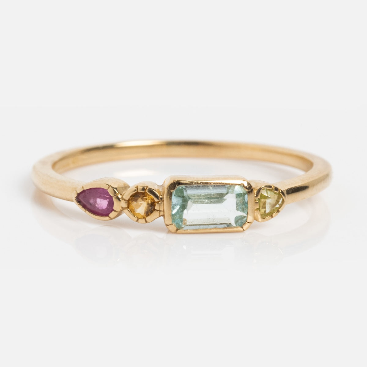 The Solid Gold First Light Pastel Gemstone Ring features a rectangular aquamarine centerpiece, with a round yellow stone, a light green stone, and a pear-shaped pink stone set in solid gold on a white background.