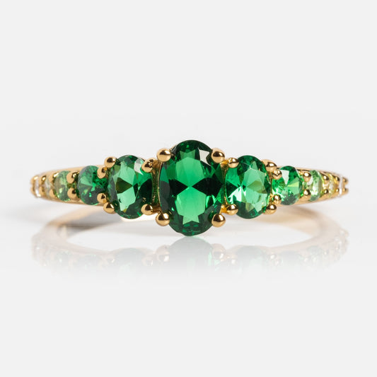 The True Colors Classic Ombre Birthstone Ring features three oval-cut green gemstones at the center and smaller green stones along a 14k gold plated band, beautifully displayed against a white background—a timeless, elegant piece.