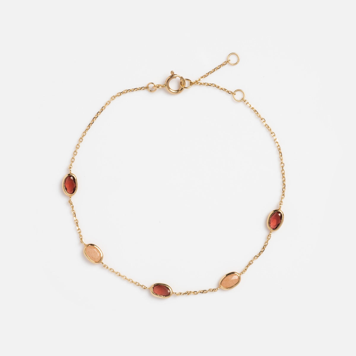 The Solid Gold Sunlit Garnet and Sunstone Bracelet features five bezel-set oval gemstones—three garnets and two sunstones—with a spring ring clasp and adjustable end. Shown on a white background.