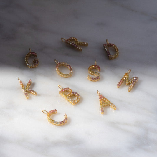 Nine Solid Gold Pastel Gemstone Letter Charms, featuring the letters C, O, S, M, R, D, K, B, and A with pastel gemstones, are arranged on a marble surface in soft natural light and delicate shadows.