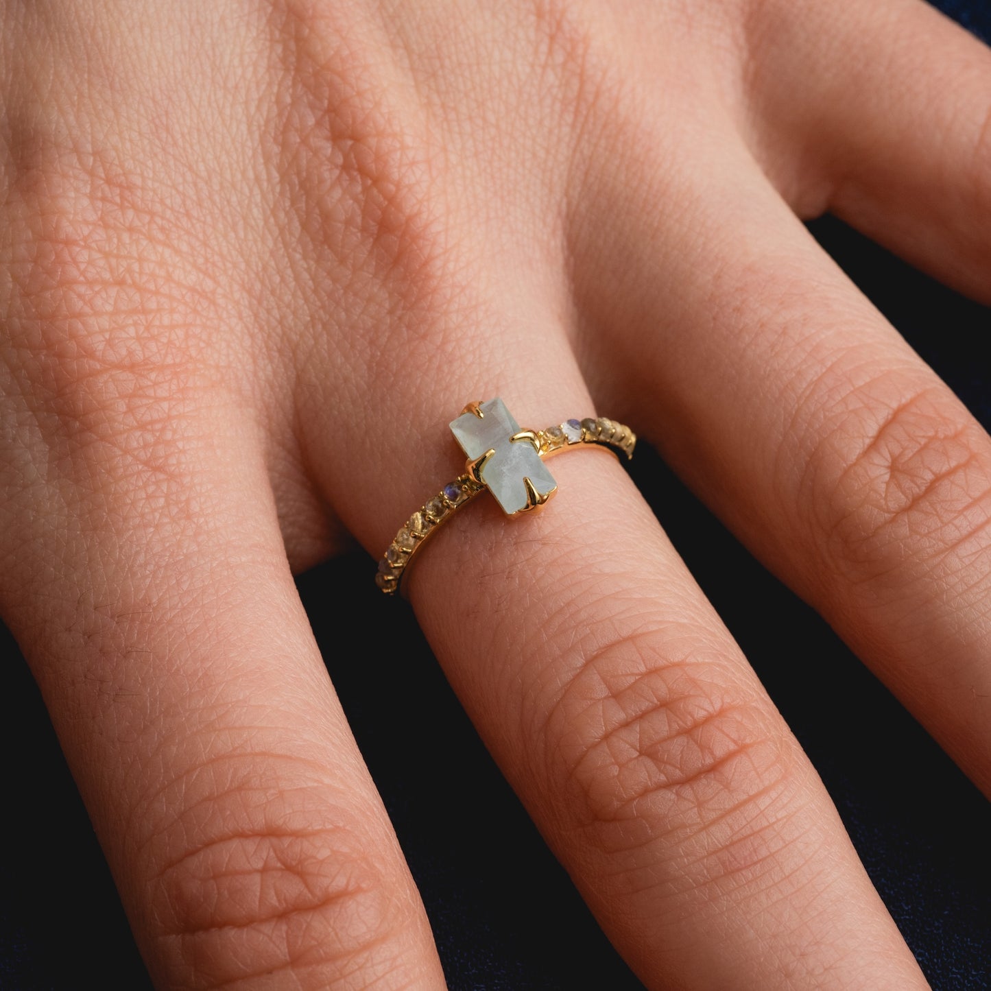 A close-up of a hand wearing the 2026 March Capsule Delicate Aquamarine Solitaire Ring, featuring a cross-shaped aquamarine center stone and small stones set along the gold band.