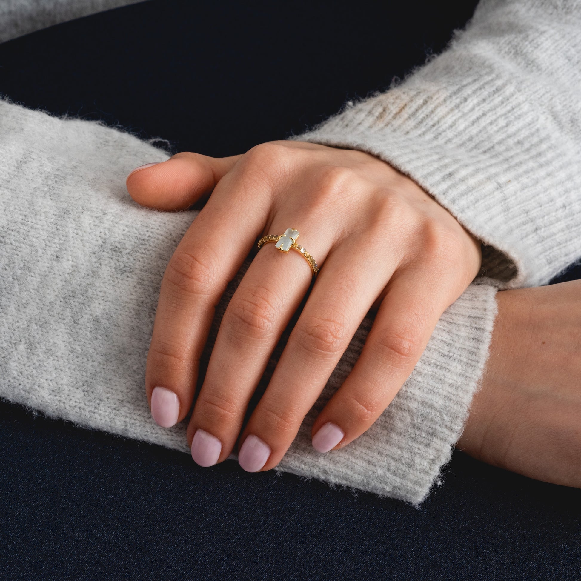 A hand with light pink nail polish rests on an arm, wearing the 2026 March Capsule Delicate Aquamarine Solitaire Ring. The person is in a light gray long-sleeve sweater, set against a dark blue background.
