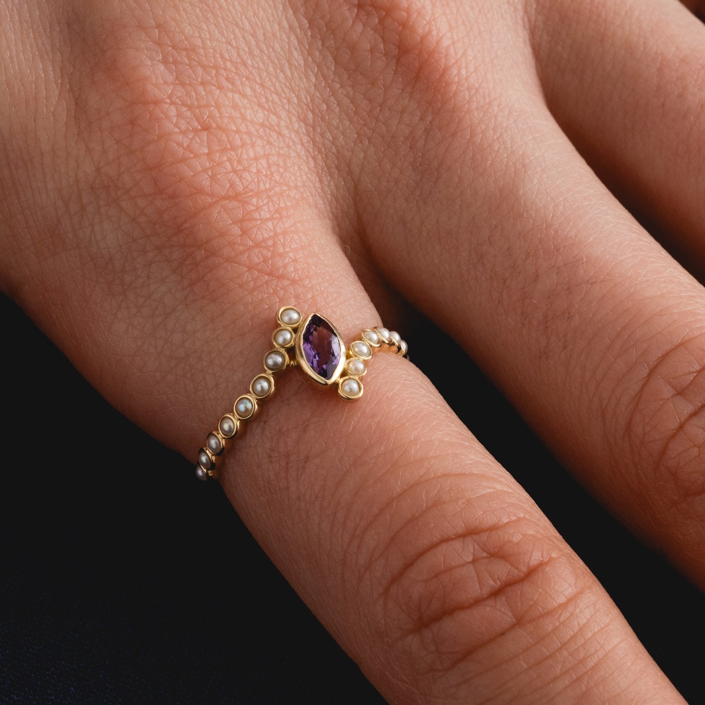 A hand wears the Solid Gold 2026 February Capsule Floating Amethyst and Pearl Twist Ring, featuring a central oval amethyst and small pearls, showcased on the middle finger against a dark background.