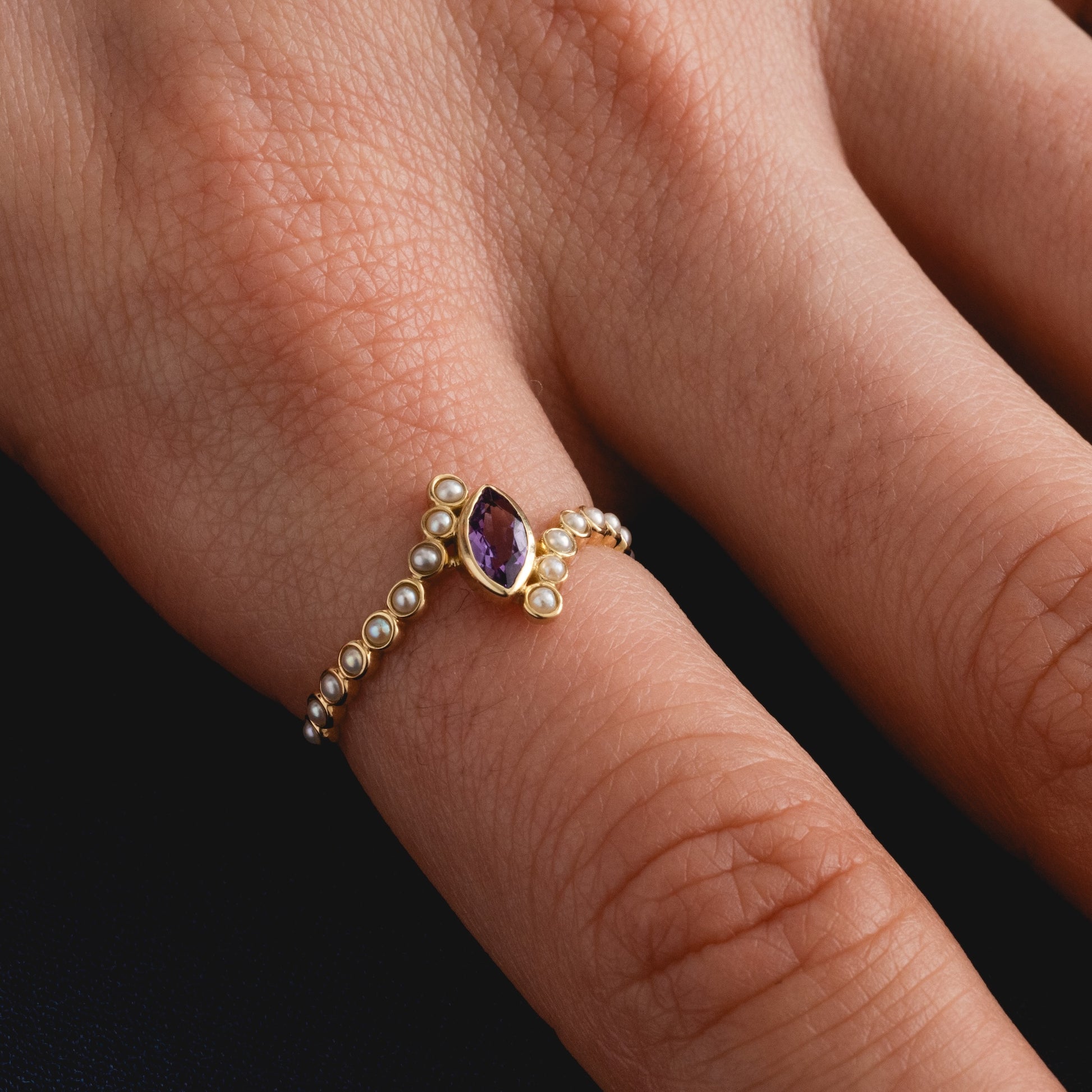 A hand wears the Solid Gold 2026 February Capsule Floating Amethyst and Pearl Twist Ring, featuring a central oval amethyst and small pearls, showcased on the middle finger against a dark background.