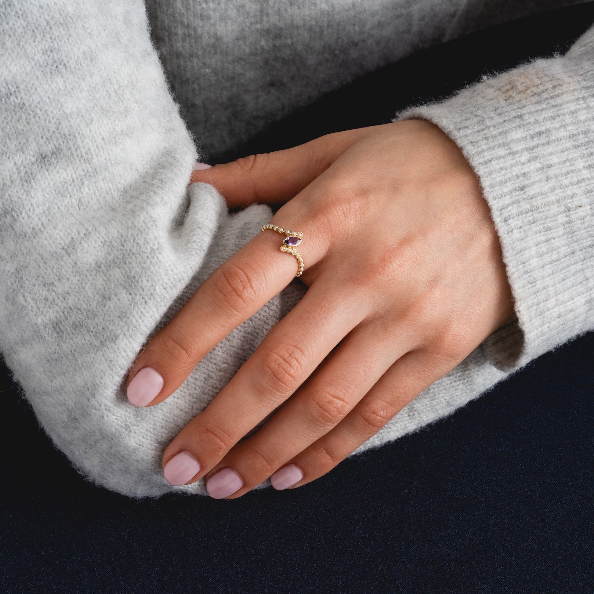 A person wearing a light gray sweater rests their hand on a dark surface, showcasing the Solid Gold 2026 February Capsule Floating Amethyst and Pearl Twist Ring on their ring finger. Their nails are neatly manicured with light pink polish.